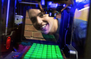 Michael Iseman looking through a 3D printer at some of the game pieces it produces for the prototype of a board game he is promoting through a $10,000 Kickstarter funding campaign.