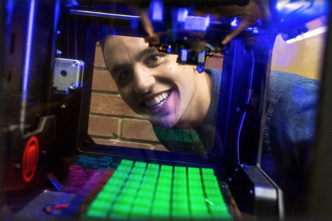 Michael Iseman looking through a 3D printer at some of the game pieces it produces for the prototype of a board game he is promoting through a $10,000 Kickstarter funding campaign.