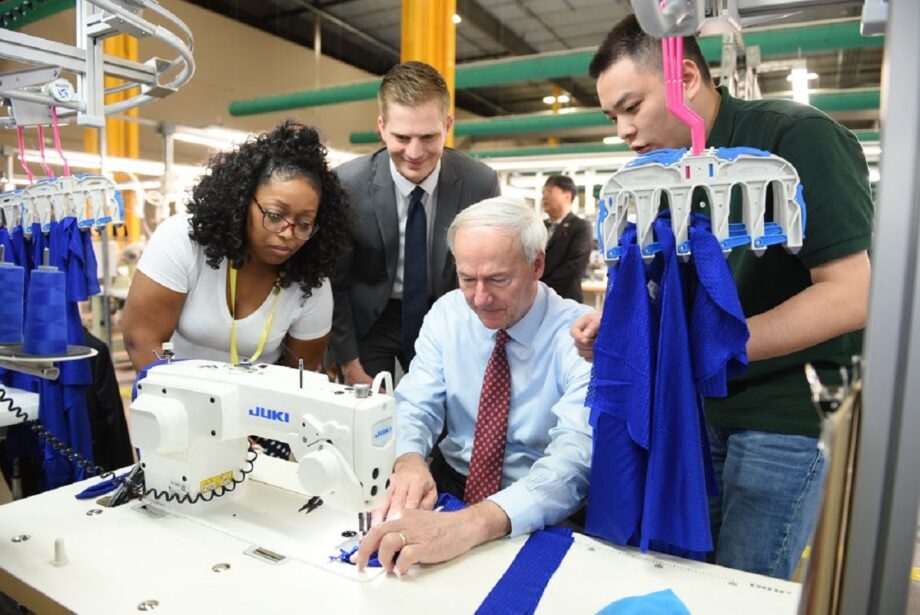 Gov. Asa Hutchinson tries his hand at sewing with a group of employees at TY Garments at their new textile and apparel facility in Little Rock.