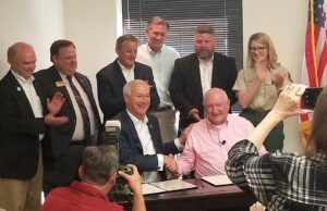 Gov. Asa Hutchinson and U.S. Secretary of Agriculture Sonny Perdue (seated) shake hands and pose for photographs in Little Rock. U.S. Reps. Bruce Westerman, French Hill, Rick Crawford and others look on.