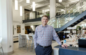 A man in a button down stands inside a modern building, leaning on the countertop of a coffee shop