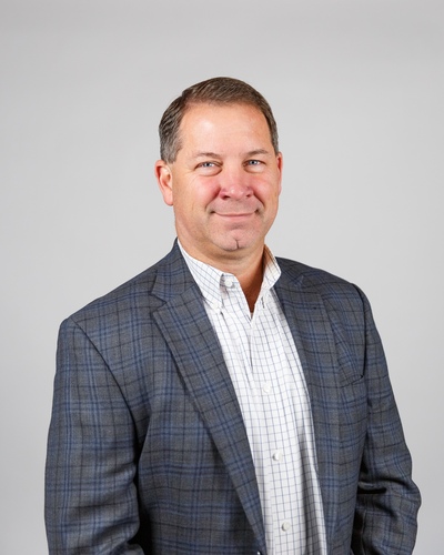 Alex Maynard, caucasian male in a dark gray suit and white collar shirt with no tie on a light gray background. 