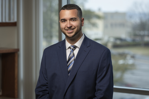 Antonije Lazic, a man with short cropped dark hair and a minimal beard wearing a dark blue suit with a striped tie on a blurred office background. 