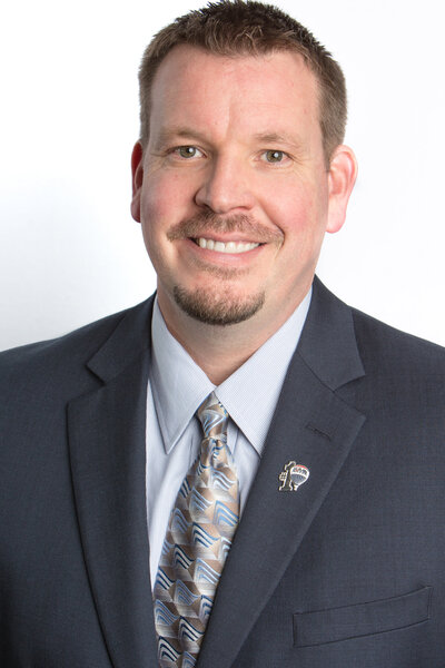 Daniel Gray, a caucasian man with a mustache and goatee in a formal navy suit with a beige and blue tie on a white background