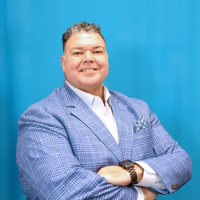 David Fox, a broad-shouldered man crossing his arms with salt and pepper curly close cropped hair wearing a light blue gingham suit with a white collared shirt. 