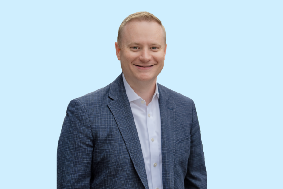Headshot of Dennis Anderson, caucasian male in a navy suit on a light blue background.