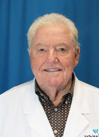 Dr. Joe Forney, an elderly white man wearing a hospital lab coat and a brown patterned collared shirt in front of a blue background. 