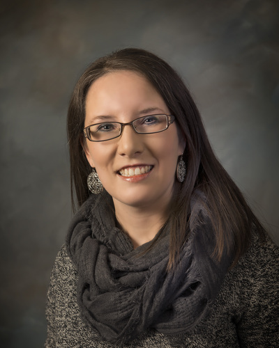 Leah DiPietro, a white woman with straight brown hair in a sweater with a black infinity scarf, thin wire-frame glasses and round dangling earrings. 