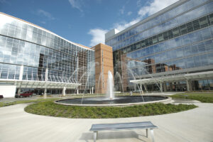 A multi-story glass and brick building sits behind a large fountain with greenery and a bench