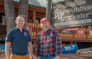 Austin Albers standing to the left of Mike Mills in front of the Buffalo Outdoor Center, a log-cabin structure retail space.