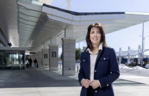 Suzanne Peyton, an older brunette woman standing outside the departure terminal of Clinton National Airport, a long decorative breezeway.