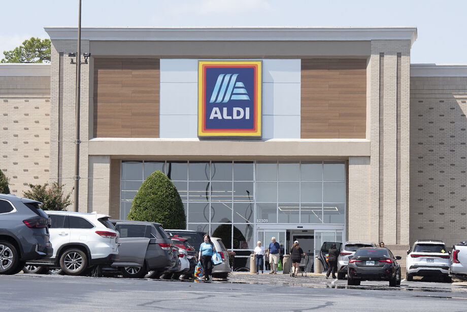 A brick building with glass windows and the Aldi logo, with a parking lot and shoppers in the foreground