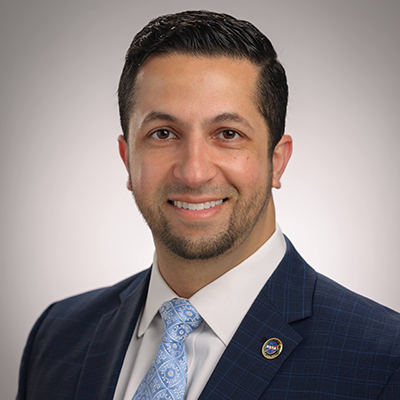 A headshot of a man with dark brown hair in a navy blue suit and light blue tie