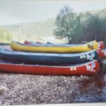 An old photograph of stacks of colorful canoes by a river.