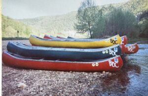 An old photograph of stacks of colorful canoes by a river.