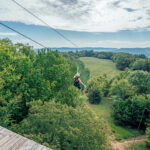 A person zip lining across a tree-filled landscape to a clearing.
