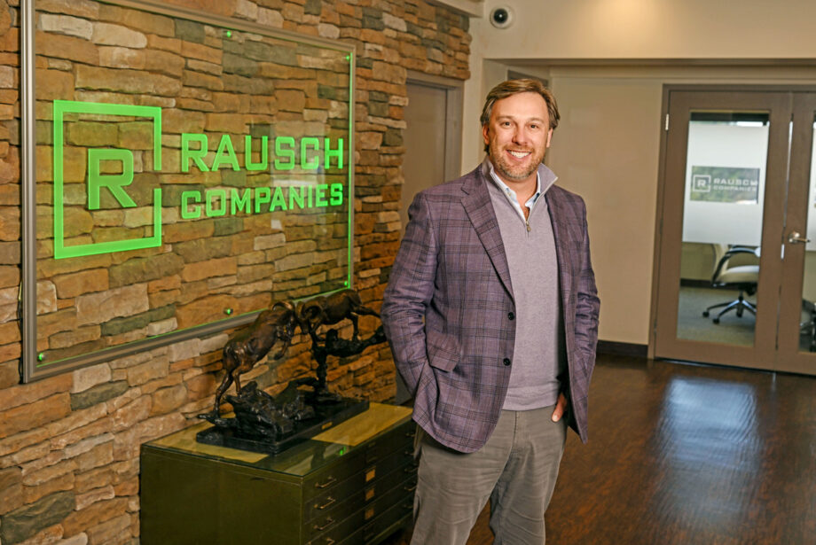 John Rausch, a middle-aged white man in a purple blazer, stands in the lobby of Rausch Companies, the logo on a sign behind him.