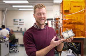 A man in a red sweater stands in an industrial engineering lab while holding a piece of silver packaging equipment
