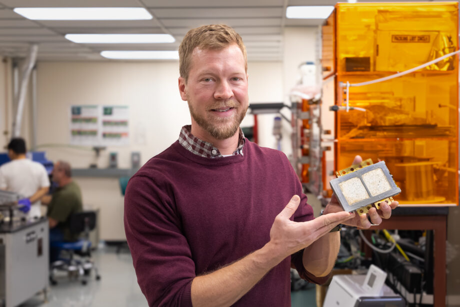 A man in a red sweater stands in an industrial engineering lab while holding a piece of silver packaging equipment