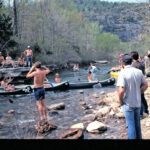 An old photo of a crowd of people canoeing and hanging around a river.