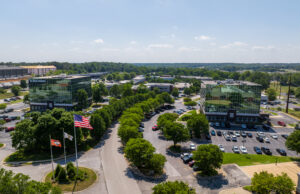 Two multistory glass office buildings sit across a road, with three flags and a parking lot in the foreground