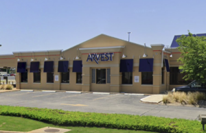 The streetview of an Arvest Bank branch, a one-story building with beige walls and blue window shutters.