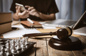 A female lawyer working on her phone and laptop on a desk with a chess set, stacks of books and journals and a gavel.