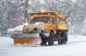 A yellow snow plow scrapes snow off a snow-covered road.