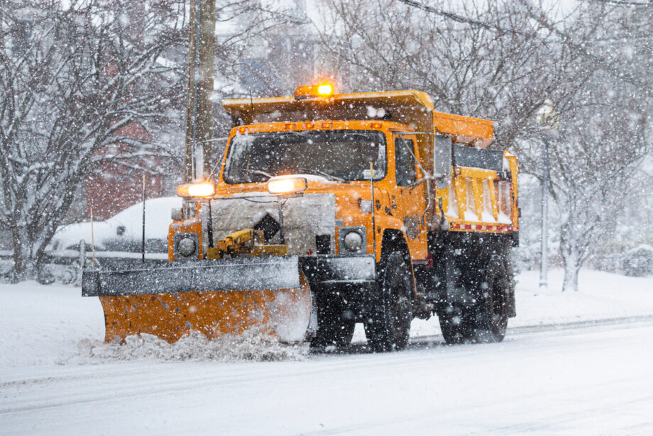 A yellow snow plow scrapes snow off a snow-covered road.