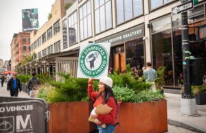 A woman in a red sweater holds a picket sign for Starbucks Workers United outside a Starbucks location