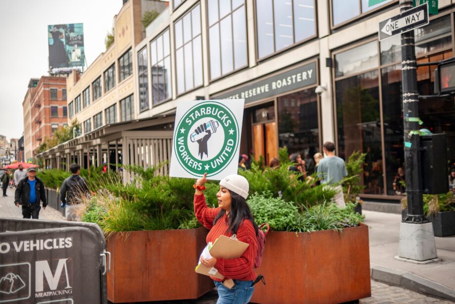 A woman in a red sweater holds a picket sign for Starbucks Workers United outside a Starbucks location