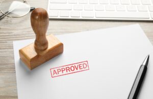 A piece of paper with an "approved" stamp next to a keyboard and a pen.
