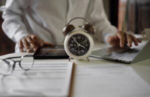 Older man at a desk with scatted paperwork, glasses and a tablet. He is working on a laptop. There is an old-fashioned alarm clock that is in the center of the frame.