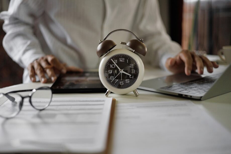Older man at a desk with scatted paperwork, glasses and a tablet. He is working on a laptop. There is an old-fashioned alarm clock that is in the center of the frame.