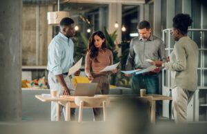 A diverse group of office workers gathered around a desk with paperwork in their hands
