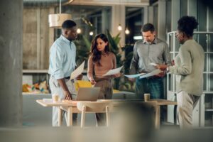 A diverse group of office workers gathered around a desk with paperwork in their hands