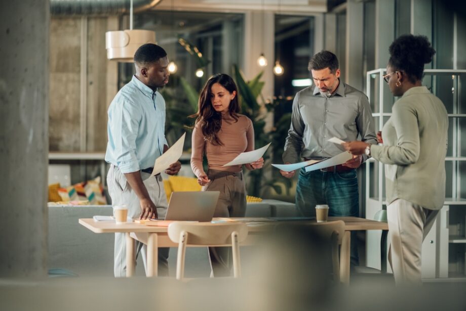 A diverse group of office workers gathered around a desk with paperwork in their hands