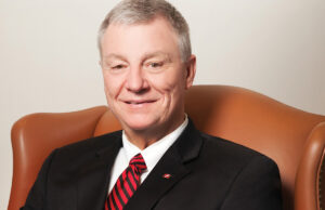 A man in a suit and red tie sits in a light brown leather chair in front of a beige wall